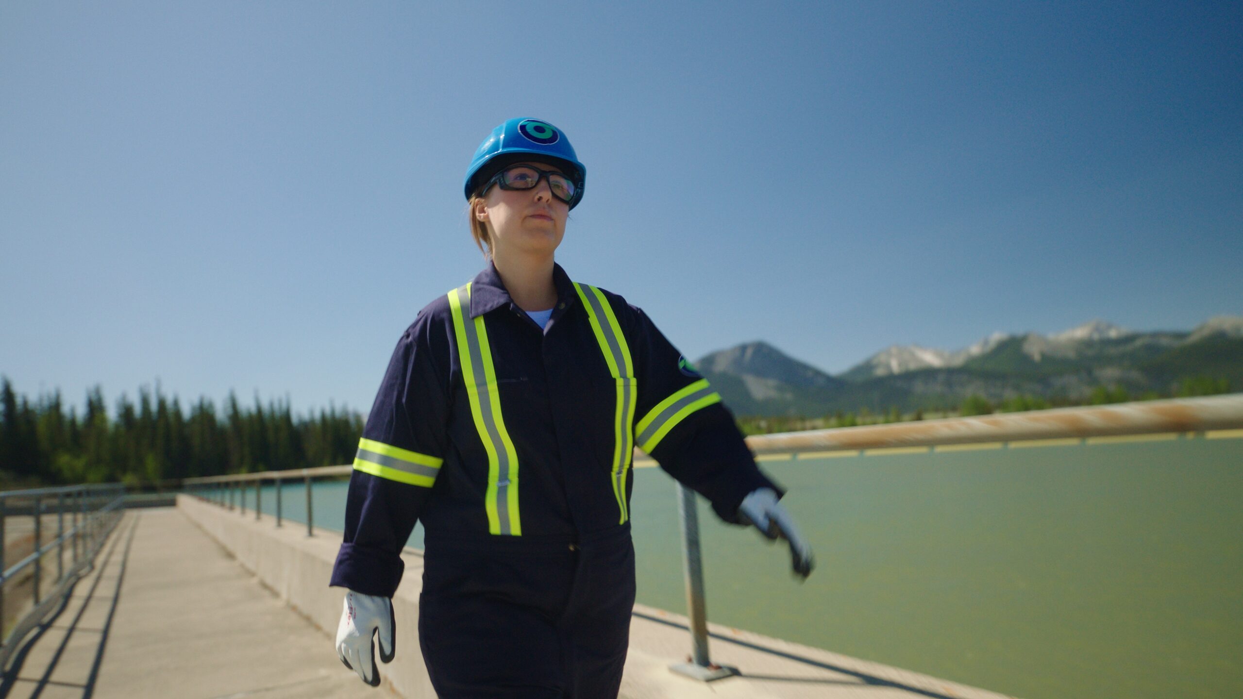 Worker in safety gear walking along a hydro facility dam, with mountains and water in the background.